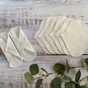 May include: A set of reusable cloth pads in a natural colour with white trim. The pads are folded and arranged on a white wooden surface. There are also some green leaves in the image.