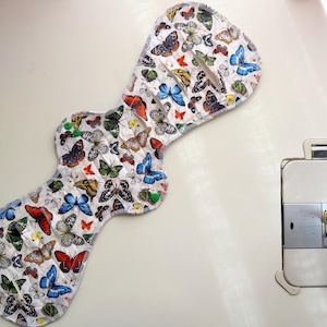 May include: A reusable cloth menstrual pad featuring a white fabric base with a colorful butterfly print in shades of blue, orange, yellow, and black. The pad has a contoured shape with two green plastic snap fasteners and visible stitching.