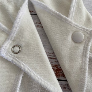 May include: Two white cloth pads with poppers and a sewn-in edge. One pad has a metal ring, the other has a white popper.