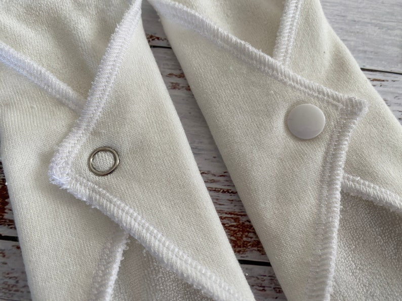May include: Two white cloth pads with poppers and a sewn-in edge. One pad has a metal ring, the other has a white popper.