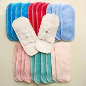 K&ouml;nnte beinhalten: Eine Sammlung wiederverwendbarer Stoffbinden in verschiedenen Farben, darunter Blau, Rot, Rosa und T&uuml;rkis. Zwei wei&szlig;e Binden mit Knopfverschluss sind ebenfalls vorhanden. Die Binden sind ordentlich angeordnet.