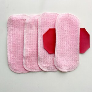 K&ouml;nnte beinhalten: Vier rosa wiederverwendbare Stoffbinden mit strukturierter Oberfl&auml;che sind auf wei&szlig;em Hintergrund angeordnet. Ein rotes, geometrisch geformtes St&uuml;ck ist an einer der Binden befestigt. Die Binden sind f&uuml;r die Damenhygiene bestimmt.