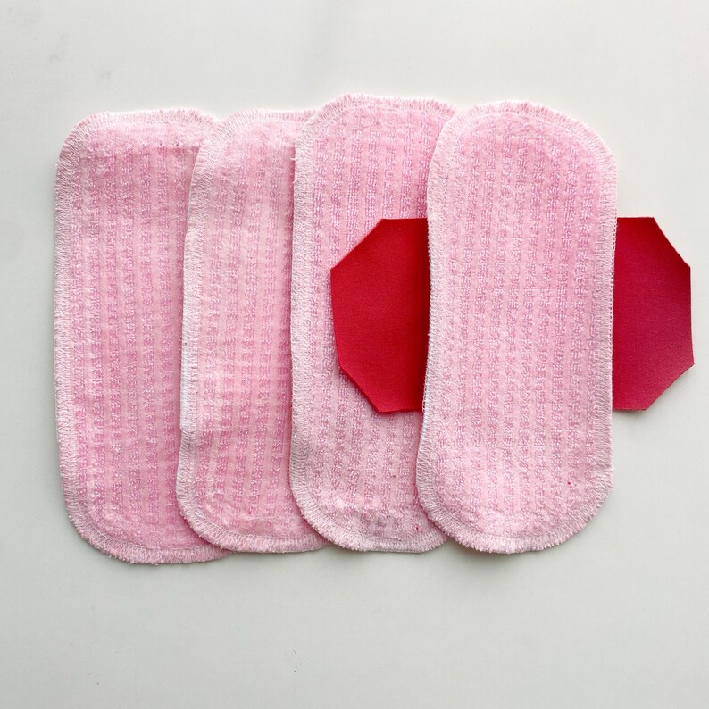 K&ouml;nnte beinhalten: Vier rosa wiederverwendbare Stoffbinden mit strukturierter Oberfl&auml;che sind auf wei&szlig;em Hintergrund angeordnet. Ein rotes, geometrisch geformtes St&uuml;ck ist an einer der Binden befestigt. Die Binden sind f&uuml;r die Damenhygiene bestimmt.