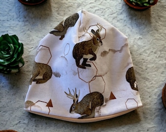 May include: White beanie with a brown rabbit and geometric pattern. The rabbit has antlers and is repeated throughout the design.