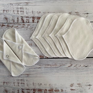 May include: A set of seven white reusable cloth pads with a white trim. The pads are folded and stacked on a white wooden surface.