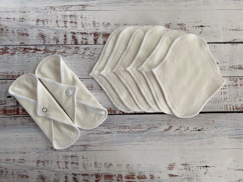 May include: A set of seven white reusable cloth pads with a white trim. The pads are folded and stacked on a white wooden surface.