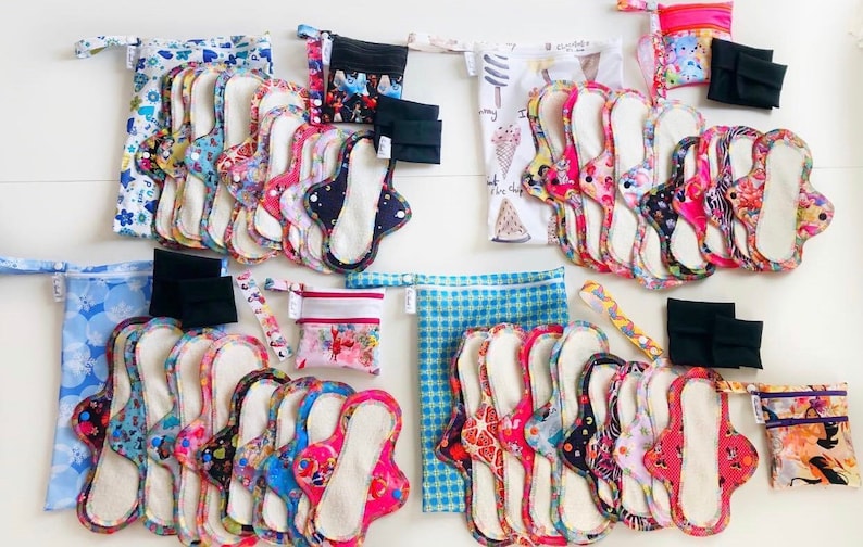 May include: A collection of reusable cloth menstrual pads in various colors and patterns, with matching storage bags. The pads are arranged in stacks and are ready to be used.
