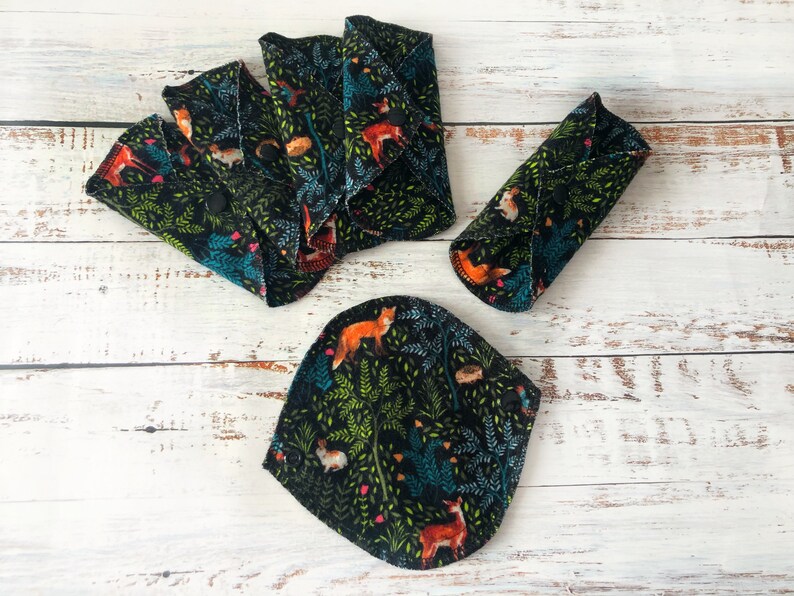 May include: Reusable cloth menstrual pads with a forest animal print. The pads feature a black background with foxes, deer, rabbits, and other forest creatures. The pads are in various shapes and sizes, with snaps for secure closure.