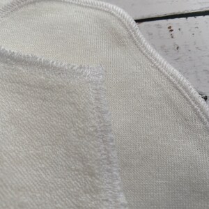 May include: Close-up of a white fabric with a stitched edge. The fabric has a soft, textured surface.