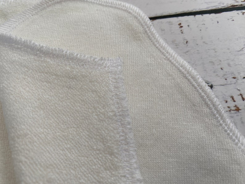May include: Close-up of a white fabric with a stitched edge. The fabric has a soft, textured surface.