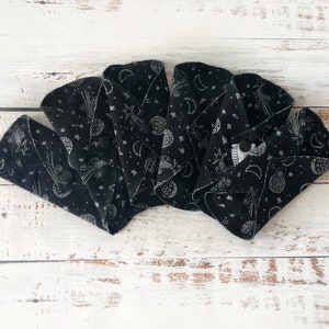 May include: Four black reusable cloth menstrual pads with a white celestial print of moons, stars, and comets. The pads are folded in half and arranged in a fan shape.