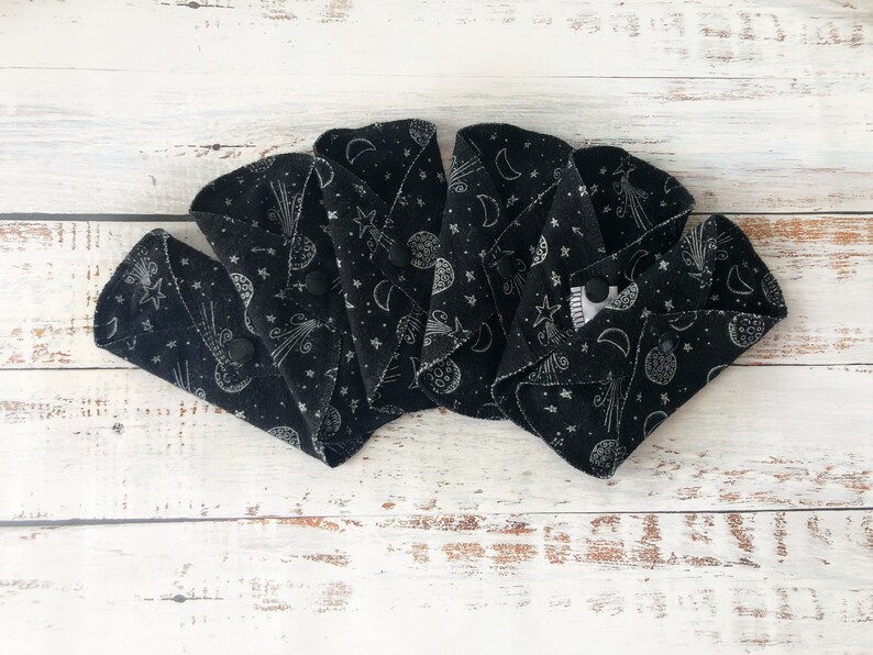 May include: Four black reusable cloth menstrual pads with a white celestial print of moons, stars, and comets. The pads are folded in half and arranged in a fan shape.