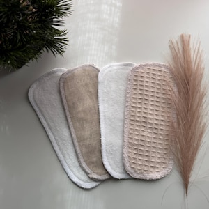 May include: Four oval-shaped reusable cloth pads arranged in a row. The set includes two white soft fabric pads, one beige linen-textured pad, and one beige waffle-weave textured pad. Each pad features a stitched border edge for durability.