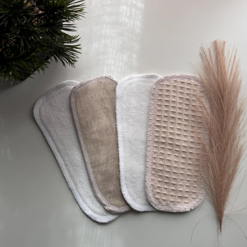 May include: Four oval-shaped reusable cloth pads arranged in a row. The set includes two white soft fabric pads, one beige linen-textured pad, and one beige waffle-weave textured pad. Each pad features a stitched border edge for durability.