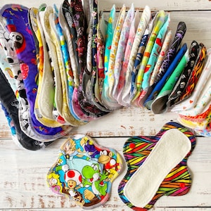May include: A collection of colorful reusable cloth pads with various patterns, including cartoon characters, animals, and abstract designs. The pads are folded and stacked on a white wooden surface.