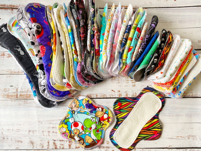 May include: A collection of colorful reusable cloth pads with various patterns, including cartoon characters, animals, and abstract designs. The pads are folded and stacked on a white wooden surface.