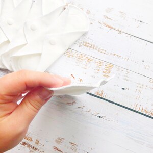 K&ouml;nnte beinhalten: Ein Stapel wei&szlig;er wiederverwendbarer Stoff-Menstruationseinlagen auf einer wei&szlig;en Holzoberfl&auml;che. Eine Hand h&auml;lt eine der Einlagen.