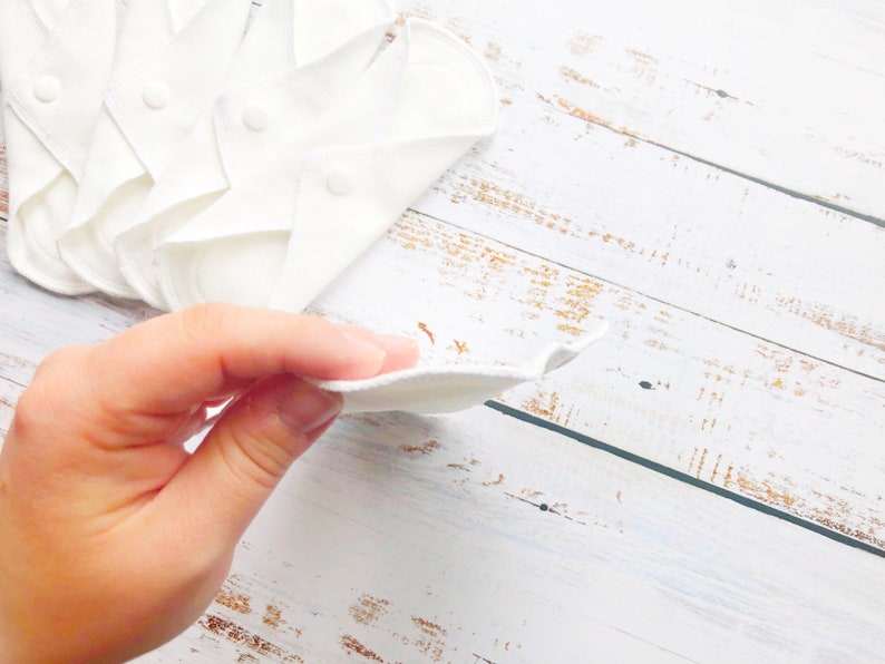 K&ouml;nnte beinhalten: Ein Stapel wei&szlig;er wiederverwendbarer Stoff-Menstruationseinlagen auf einer wei&szlig;en Holzoberfl&auml;che. Eine Hand h&auml;lt eine der Einlagen.