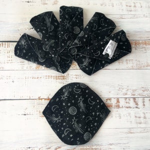 May include: A set of six black reusable cloth menstrual pads with a white star and crescent moon pattern. The pads are folded and arranged in a fan shape on a white wooden surface.