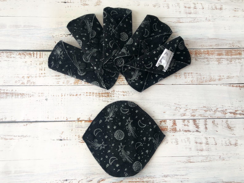 May include: A set of six black reusable cloth menstrual pads with a white star and crescent moon pattern. The pads are folded and arranged in a fan shape on a white wooden surface.