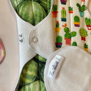 May include: A white cloth menstrual pad with a green watermelon print on one side and a cactus print on the other side. The pad has a white fleece lining and a snap closure. The pad is labeled "Eco Adorablle" and "PP".
