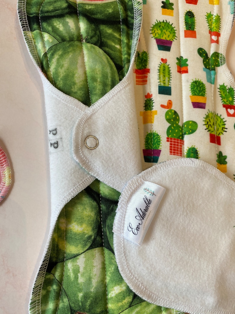 May include: A white cloth menstrual pad with a green watermelon print on one side and a cactus print on the other side. The pad has a white fleece lining and a snap closure. The pad is labeled "Eco Adorablle" and "PP".