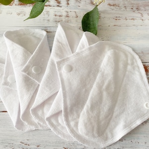 May include: Three white reusable cloth menstrual pads with snaps. The pads are folded and arranged on a white wooden surface.