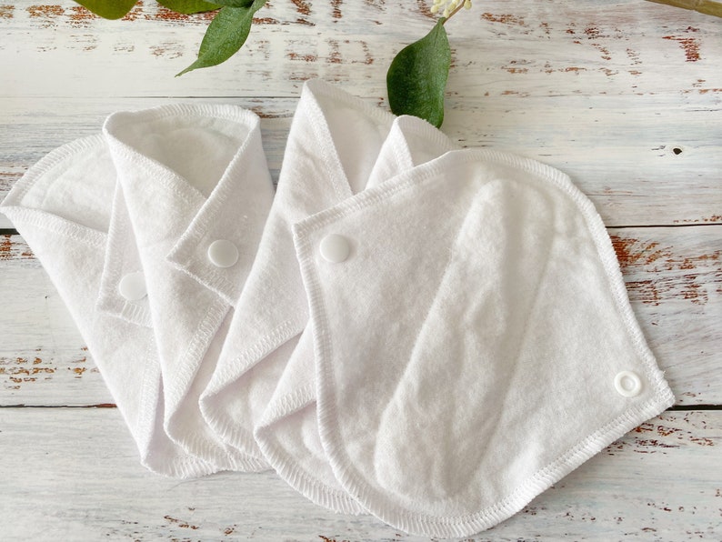 May include: Three white reusable cloth menstrual pads with snaps. The pads are folded and arranged on a white wooden surface.