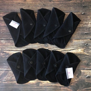 May include: Ten black reusable cloth menstrual pads with snaps. The pads are folded in half and arranged in two rows of five. The pads have a white label with black text that says "Sustainable Life".