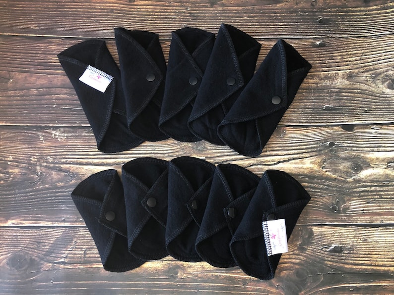 May include: Ten black reusable cloth menstrual pads with snaps. The pads are folded in half and arranged in two rows of five. The pads have a white label with black text that says "Sustainable Life".
