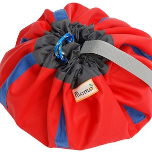 Könnte beinhalten: Eine rot-blaue Spielmatte und Aufbewahrungstasche mit Kordelzug. Die Tasche hat einen grauen Griff und blaue und graue Akzente. Auf der Tasche steht das Wort "Momo" auf einem kleinen Etikett.