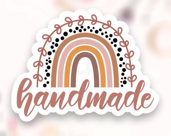 Könnte beinhalten: Ein Aufkleber mit einem Regenbogen-Design in den Farben Rosa, Orange und Braun. Der Regenbogen hat schwarze Punkte und einen Blatt-Rand. Das Wort "handgemacht" ist in einer kursivschriftlichen Schrift unter dem Regenbogen geschrieben.