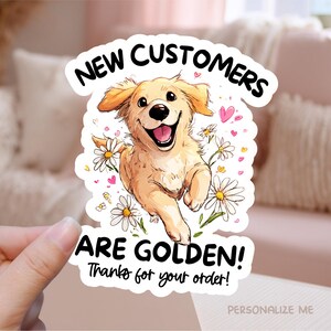 Puede incluir: Una pegatina con un perro golden retriever de dibujos animados corriendo entre margaritas. El texto dice "NEW CUSTOMERS ARE GOLDEN! Thanks for your order!" con corazones rosas. La pegatina está sujeta por una mano.