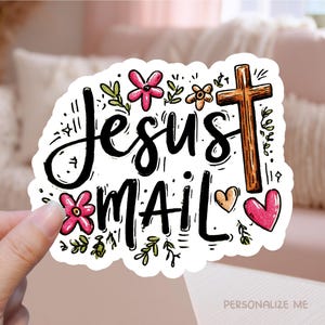 May include: A white sticker with the words "Jesus Mail" in black script, decorated with pink and orange flowers, green leaves, a brown cross, and two hearts. The sticker is held by a hand.