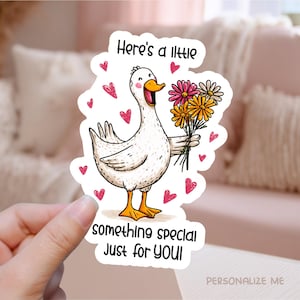 以下が含まれることがあります： 花束を持った漫画のガチョウのステッカー。ガチョウは黄色いくちばしと足を持ち、「Here's a little something special Just for YOU!」というテキストとピンクのハートが付いています。