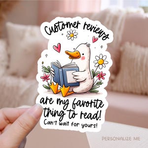 May include: A sticker featuring a cartoon duck reading a book, surrounded by flowers and hearts. The text reads "Customer reviews are my favorite thing to read! Can't wait for yours!" The sticker has a white background.