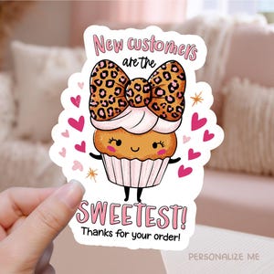 以下が含まれることがあります： ヒョウ柄のリボンが付いた漫画のカップケーキのステッカーで、「New customers are the sweetest! Thanks for your order!」という文字が入っています。ピンクのハートと星がカップケーキを囲んでいます。