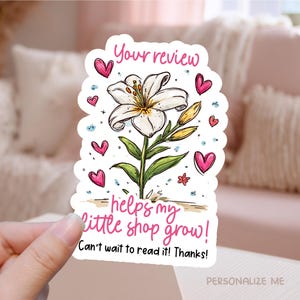 May include: A sticker featuring a white lily with green leaves and pink hearts. The text "Your review helps my little shop grow!" is written in pink script. The sticker is held by a hand.