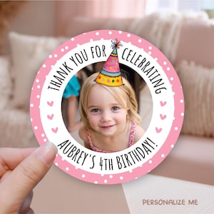 May include: A round, pink sticker with a photo of a young girl wearing a party hat. The sticker has the text "THANK YOU FOR CELEBRATING AUBREY'S 4TH BIRTHDAY!" in a circular design with white polka dots and pink hearts.