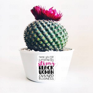May include: A white ceramic pot with a cactus plant and a pink flower on top. The pot has a sticker that says "Thank you for supporting this strong black woman owned business".