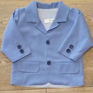 May include: A light blue blazer with a textured pattern, two front pockets, and three buttons on each sleeve. The blazer has a notched collar and three buttons down the front. The label reads "DOMINIK".