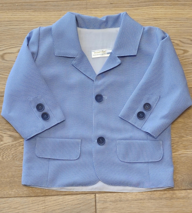 May include: A light blue blazer with a textured pattern, two front pockets, and three buttons on each sleeve. The blazer has a notched collar and three buttons down the front. The label reads "DOMINIK".