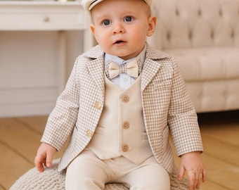 May include: A baby wearing a beige plaid blazer, vest, and pants, paired with a light blue shirt and bow tie. The outfit includes a matching beige cap. The baby is sitting on a textured, light-colored ottoman.