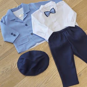 May include: A light blue blazer, white button-down shirt with a navy bow tie, navy pants, and a navy cap. The children's formal wear set is laid out on a wooden surface.