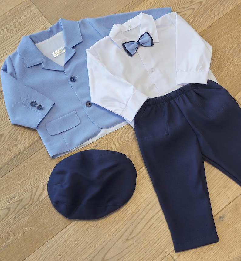 May include: A light blue blazer, white button-down shirt with a navy bow tie, navy pants, and a navy cap. The children's formal wear set is laid out on a wooden surface.