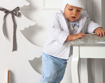 May include: A young child wearing a white quilted jacket, light blue pants, and a white hat. The child is standing next to a white wooden stool and a white paper Christmas tree.