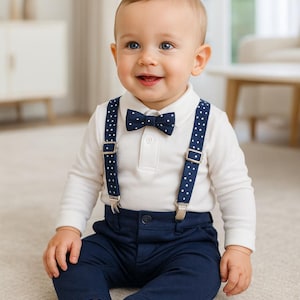 Op de afbeelding: Een baby in een outfit met een wit shirt met lange mouwen, een marineblauwe broek en een marineblauwe vlinderdas met witte stippen. De outfit bevat bijpassende bretels met stippen. De baby zit op een lichtgekleurd tapijt.
