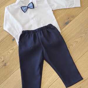 May include: A white long-sleeved shirt with a blue bow tie and navy blue pants. The shirt has a button-down front and a collar. The pants have an elastic waistband and pockets. This outfit is suitable for formal occasions.