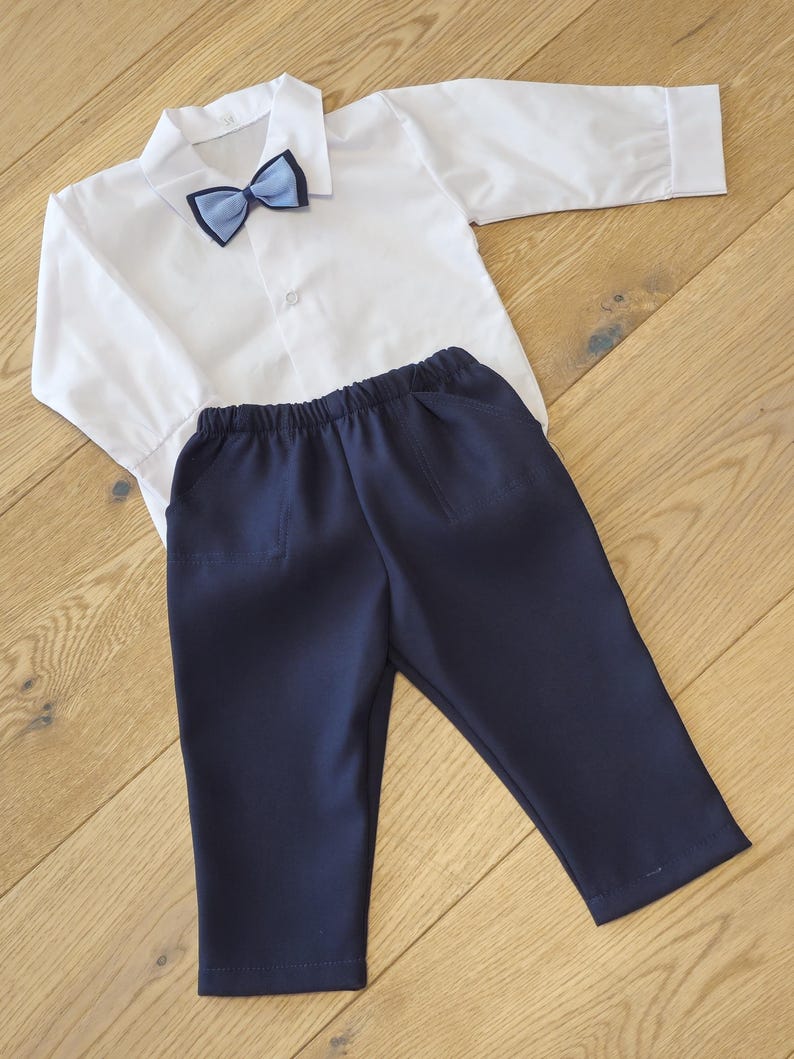 May include: A white long-sleeved shirt with a blue bow tie and navy blue pants. The shirt has a button-down front and a collar. The pants have an elastic waistband and pockets. This outfit is suitable for formal occasions.