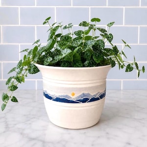 May include: A cream-colored ceramic planter with a flared rim, featuring a hand-painted landscape band around the center depicting blue mountain silhouettes, a light gray sky, and a small yellow sun. A trailing green plant with variegated leaves.